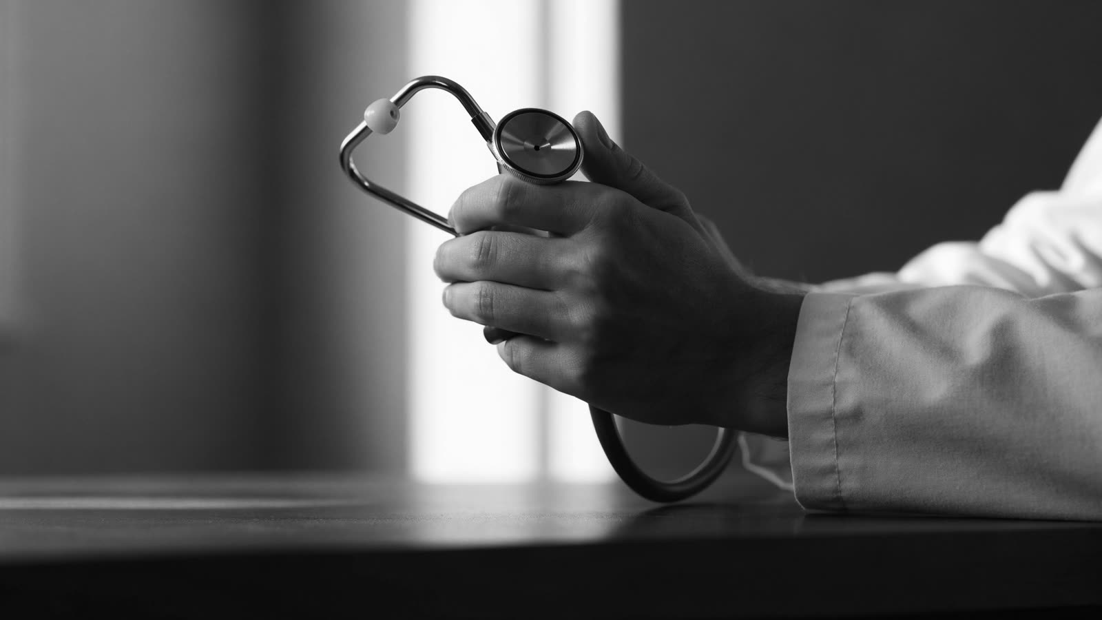 Mãos de médico segurando um estetoscópio sobre mesa escura, em preto e branco