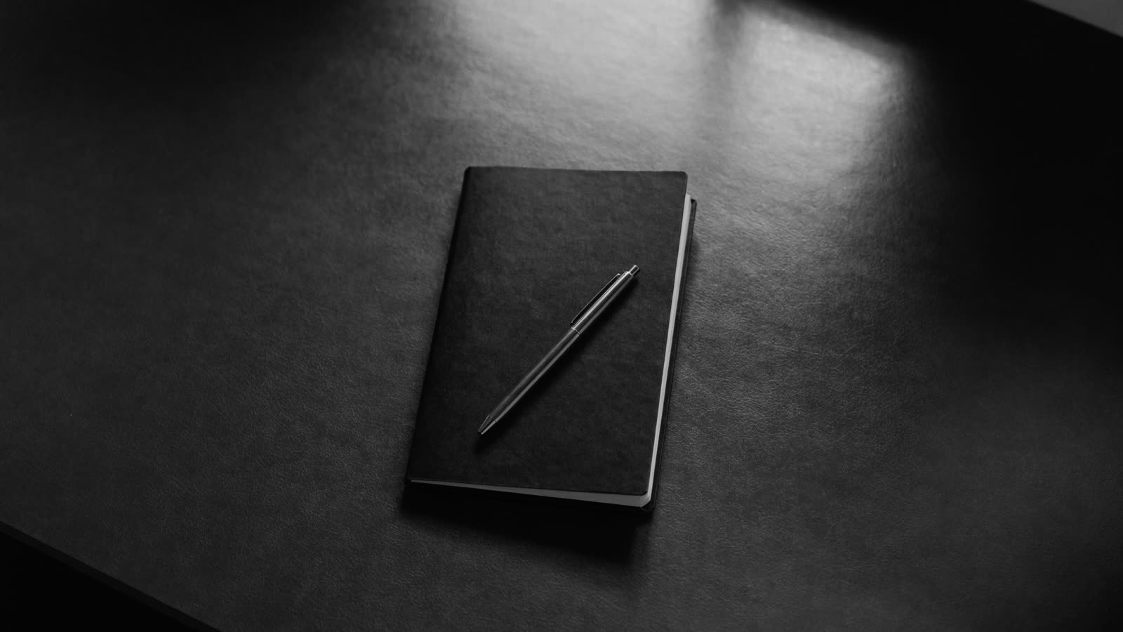 Caderno preto fechado e uma caneta, vistos de cima sobre mesa escura
